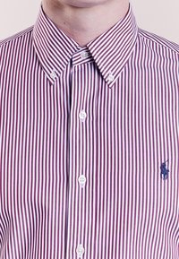 Randig skjorta med vertikala röda och vita ränder, knäppning i kragen, vita knappar och en blå broderad logotyp på bröstet.