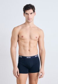Boxeri scurți de culoare bleu marin, confecționați dintr-un material moale, având un brad cu cuvântul "BOSS" scris cu alb. Textură netedă, design ajustat.