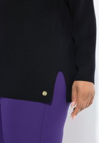 Rechterhand van een persoon die paarse broek en een zwarte trui draagt met een klein rond gouden embleem nabij de zoom.