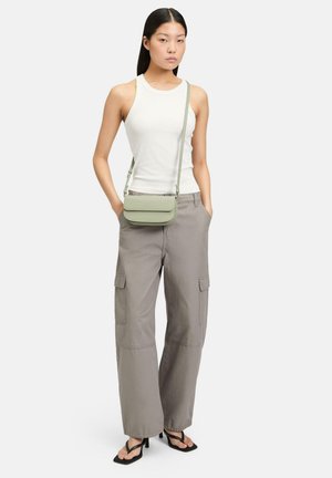 Sac bandoulière vert clair avec une texture lisse, de forme rectangulaire, et un rabat, associé à un pantalon cargo gris à jambes larges et un débardeur blanc.