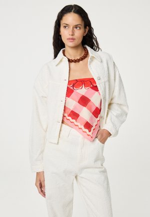 Frau trägt eine strukturierte weiße Jacke und Hose mit einem rot-rosa gemusterten Tuch als Oberteil und einer braunen Perlenkette, steht mit der Hand in der Tasche.