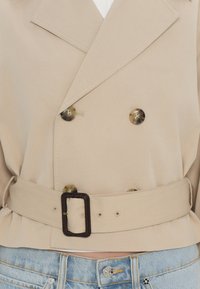 Veste beige à double boutonnage avec boutons en écaille de tortue et large ceinture à boucle rectangulaire foncée, portée sur un jean en denim bleu clair.