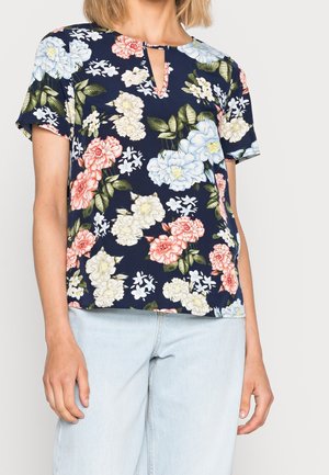 Kvinde iført en marineblå bluse med korte ærmer med lyserødt, hvidt og blåt blomsterprint og lyseblå jeans.