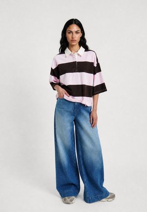 Woman wearing loose wide-leg blue jeans and a pink and black broad striped rugby shirt with white collar, standing against a plain background.