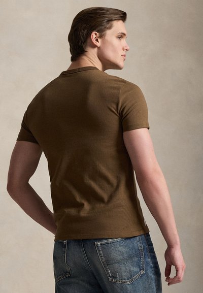 Mlad moški s kratkimi rjavimi lasmi, oblečen v oprijeto rjavo majico s kratkimi rokavi in modre kavbojke, stoji v profilu na enostavnem ozadju.