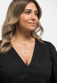 Collana in oro con cinque perle bianche, con una goccia di perla a forma di pera più grande all'estremità. Sullo sfondo di una maglietta nera.