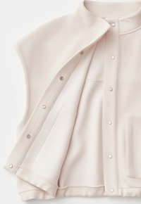 Gilet senza maniche color crema con bottoni a pressione e cuciture interne visibili, disteso in piano e parzialmente aperto su uno sfondo bianco.