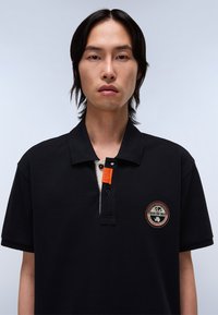 Svart polo-shirt med krage, med en kontrasterande tan placket och en orange detalj. Logotyp patch på vänster bröst.