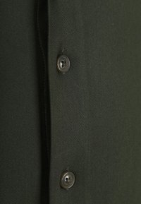 Dunkelgrüner Stoff mit glatter Textur, versehen mit einem Knopfverschluss aus dunklem Kunststoff im Vierlochdesign.