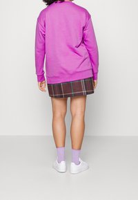 Person klädd i en lila sweatshirt, rutiga kortbyxor, lavendelfärgade strumpor och vita sneakers, stående mot en enkel ljus bakgrund.