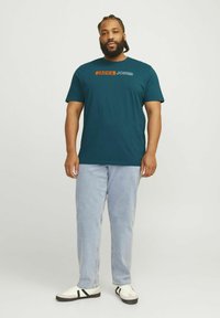 Teal bomulds t-shirt med orange og hvid "JACK & JONES" logo, parret med lyseblå løse jeans og hvide sneakers med sorte detaljer.