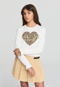 Biała koszula z długim rękawem z graficznym wzorem serca w panterkę, z złotym napisem "WIEŻ w siebie." Połączona z jasnobeżową plisowaną spódnicą.