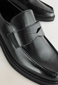 Schwarze Lederloafers mit einer glatten, glänzenden Oberfläche, ausgestattet mit einem Riemendetail und sichtbaren Nähten entlang der Sohle.