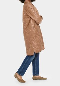 Vêtements d'extérieur matelassés marron avec une coupe décontractée, dotés de manches longues, de poches latérales et de boutons décoratifs. Associés à un jean bleu et des chaussures plates.