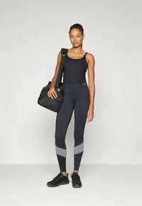 Czarny sportowy top na ramiączkach i granatowe legginsy z szarymi akcentami. Model trzyma czarną torbę sportową, ma na sobie czarne sneakersy. Prosty, dopasowany design.