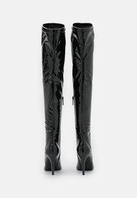 Botas de charol negras hasta el muslo con punteras afiladas, tacones de aguja y cremalleras en la parte trasera, mostradas desde atrás sobre un fondo blanco.