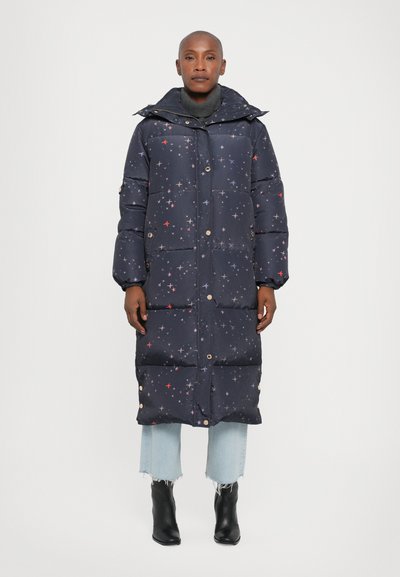 Marineblå pufferjakke med stjernemønster, lang model, trykknapper og hætte. Båret over lyseblå jeans og ankelstøvler med spids tå.
