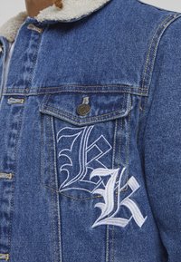 Denim jakna ima ovratnik podstavljen s sherpa materijalom, dve prsne žepove i istaknuti beli vezeni natpis na levoj strani. Modra barva in teksturiran material.
