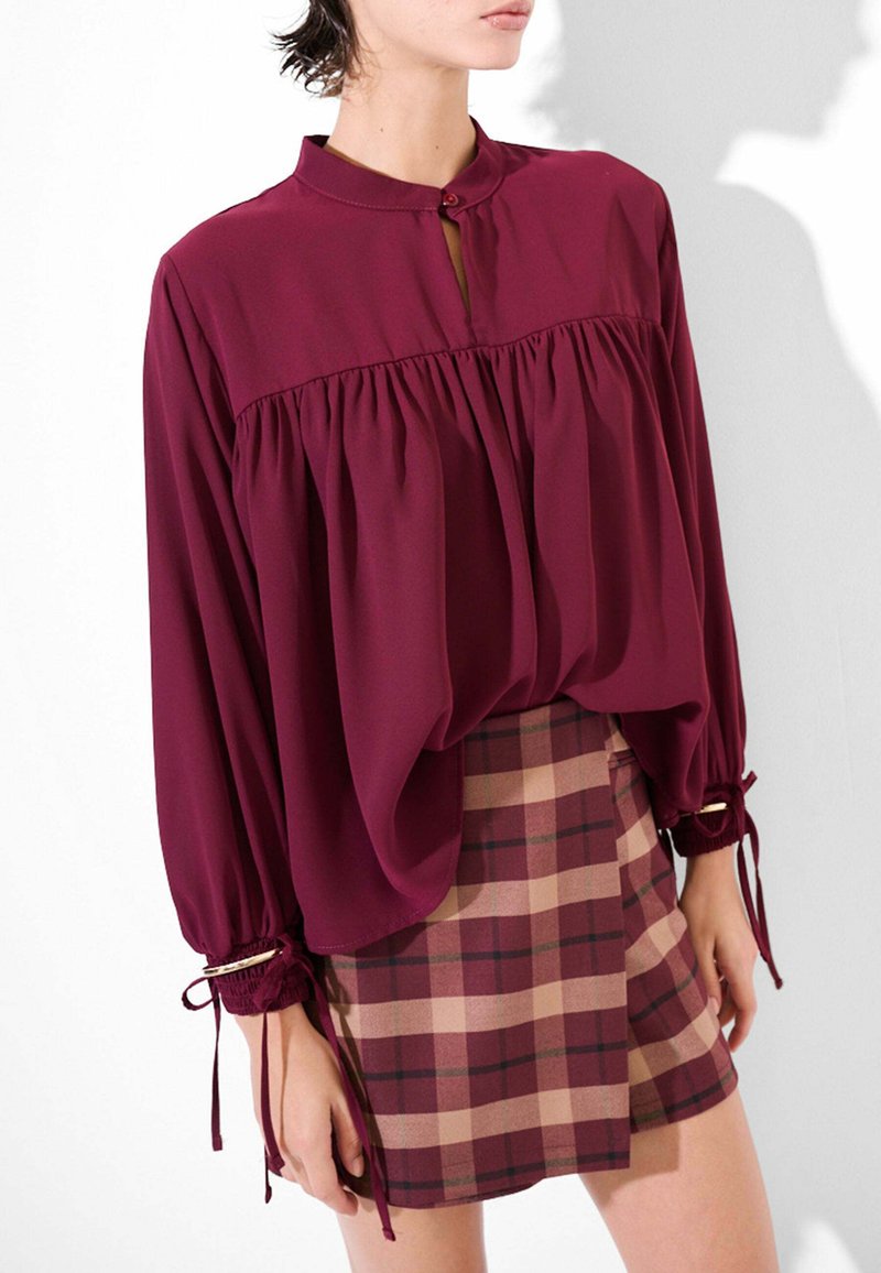 Blusa bordeaux con colletto alla mandarino, spalle arricciate e maniche a sbuffo. Abbinata a una mini gonna a quadretti in tonalità bordeaux e beige.