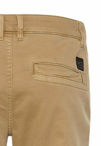 Hellbraune Hosen mit sichtbaren Nähten und einer Gesäßtasche, die ein kleines schwarzes Logo am Taillenbund trägt.