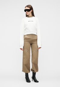 Mujer con sudadera blanca de gran tamaño, pantalones de tiro alto color beige con bolsillos con cremallera, botas negras de tacón y grandes gafas de sol negras.