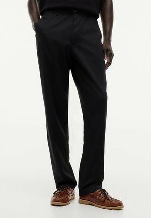 Mężczyzna w czarnych spodniach i brązowych skórzanych sznurowanych butach, stojący z rękami w kieszeniach na jednolitym tle.