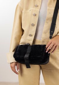 Črna torbica iz umetnega krzna z teksturirano površino, zaprta s črnim trakom in zaponko. Oseba v bež obleki.