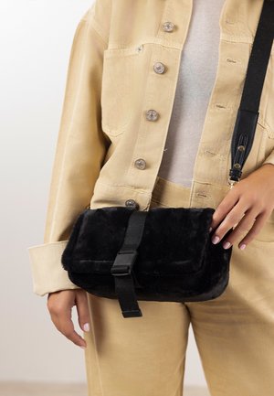 Sort skuldertaske i imiteret pels med en tekstureret overflade, sikret med en sort rem og spænde. Person iført beige outfit.