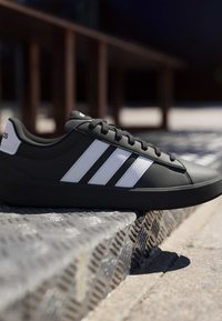 Baskets Adidas noires avec des bandes blanches et des lacets reposant sur une marche en métal texturé en plein air au soleil.