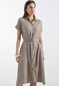 Una donna indossa un vestito camicia beige con maniche arrotolate e una vita cintata, abbinato a un bracciale spesso, su uno sfondo semplice.