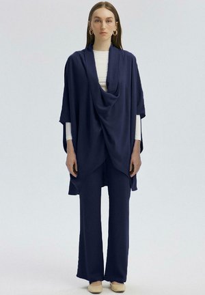 Navy draped poncho over a white long-sleeve top, paired with matching wide-leg trousers. Smooth fabric with subtle texture, earthy tone shoes.