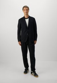 Mand står iført en sort habit med en hvid skjorte og sorte Adidas sneakers med hvide striber foran en ensfarvet baggrund.