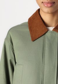 Jachetă de culoare verde măslin cu guler din catifea maro, având un design curat și structurat și cusături subtile, cu o textură de țesătură fină.