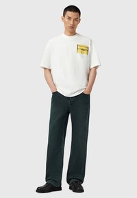Hvid bomulds-T-shirt med et gult grafisk lommeprint, parret med mørkegrønne denim wide-leg bukser og sorte sko.