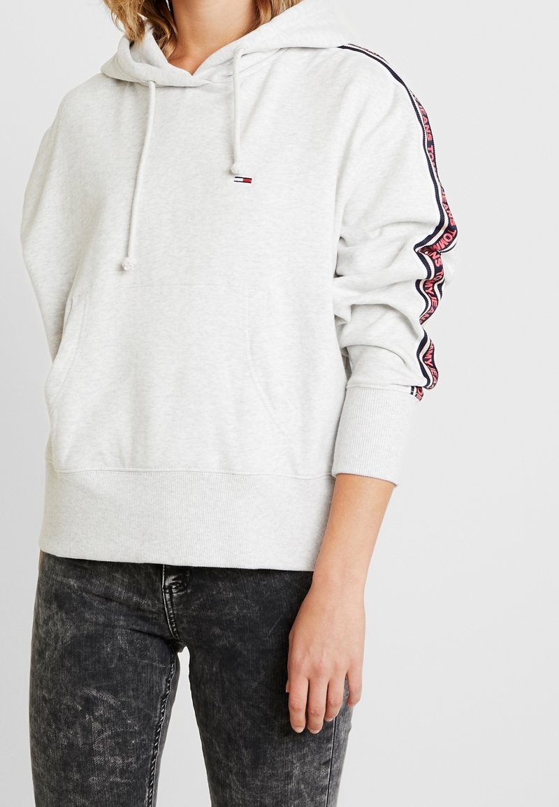 Sweat à capuche gris clair avec une poche avant, capuche à cordon de serrage, et des bandes contrastées sur les manches avec des accents rouges et bleu marine.