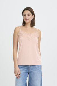 Ženska, ki nosi svetlo roza top z mrežico čipke in svetlo modre kavbojke, stoji ob belem ozadju.