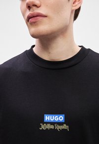 Μαύρο βαμβακε ρό μπλουζάκι με στρογγυλή λαιμόκοψη. Γραφικά περιλαμβάνουν την μπλέ ετικέτα "HUGO" και το "Jujutsu Kaisen" σε κίτρινο, τοποθετημένα κεντρικά.