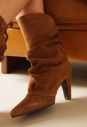 Brown suede slouchy mid-calf boots with pointed toes and high heels worn by a person sitting on a tan leather couch.