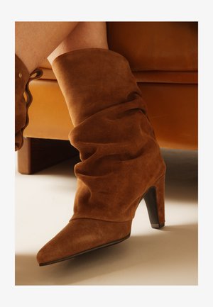 Rudos zomšos nusileidžiantys vidutinio aukščio aulinukai su smailiais noseliais ir aukštais kulnais, dėvimi žmogaus, sėdinčio ant rudos spalvos odinės sofos.