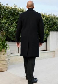 Abrigo de lana negro con cuello estructurado, mangas largas y corte recto, combinado con pantalones de raya diplomática grises y zapatos negros.