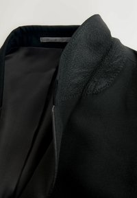 Sort jakke med en tekstureret uldkrave, glat stof og en kontrastfarvet inderfor synligt ved halsudskæringen. Rene, moderne designdetaljer.