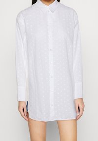 Camicia bianca a maniche lunghe con colletto, caratterizzata da un motivo goffrato a pois e chiusura frontale con bottoni.