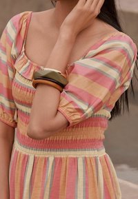 Femme portant une robe ray ée rose, jaune et grise, avec des manches bouffantes et trois bracelets en chunky à son poignet.