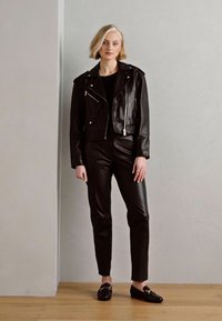 Zwarte leren motorjack met ritssluitingdetails, gecombineerd met strakke zwarte leren broek en zwarte instappers, op een houten vloer.