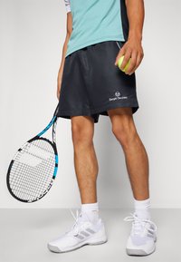 Schwarze Sportshorts mit einem Logo, kombiniert mit weißen Tennisschuhen und weißen Socken. In einer Hand wird ein Tennisschläger und ein grüner Ball gehalten.