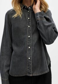 Chemise en denim gris avec une texture douce, fermeture à boutons et boutons blancs. Elle possède des manches longues et des coutures contrastées le long des bords.