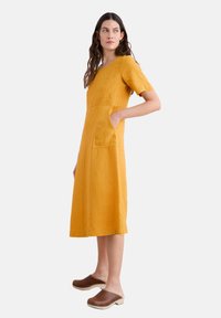 Hořčicově žluté lněné šaty s krátkými rukávy, do délky po kolena, s čtvercovým dekoltem a dvěma bočními kapsami. V kombinaci s hnědými klíny.