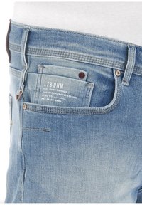 Jeans de mezclilla azul claro con un diseño de múltiples bolsillos, costuras visibles y un acento de cuero marrón en la cinturilla. Etiqueta visible en el bolsillo.