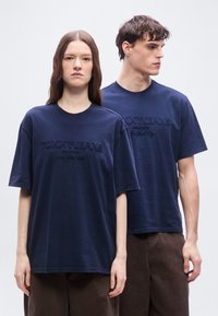 Δύο νέοι ενήλικες που φοράνε μπλε ναυτικό μπλουζάκια Tommy Jeans και καφέ παντελόνια, στέκονται μπροστά σε ένα απλό φωτεινό φόντο.