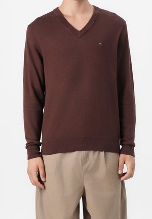 Mand iført en brun V-hals sweater med et lille logo på brystet og beige bukser mod en hvid baggrund.